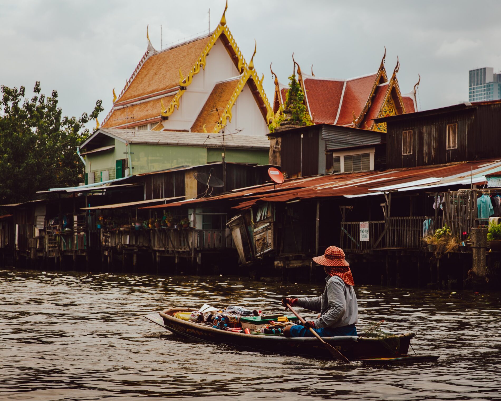 Thai woman rowing boat in front of riverside shops, homes and temples, Bangkok, Thailand, November 2017