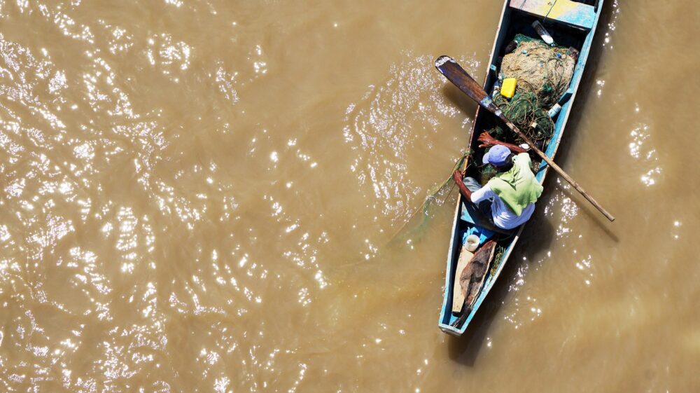 Man sitting on blue fishing rowboat in muddy water taken from above, Guayaquil, Ecuador