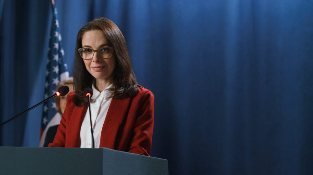 Female politician in red jacket smiling at camera standing at lecturn in front of blue curtain and US flag