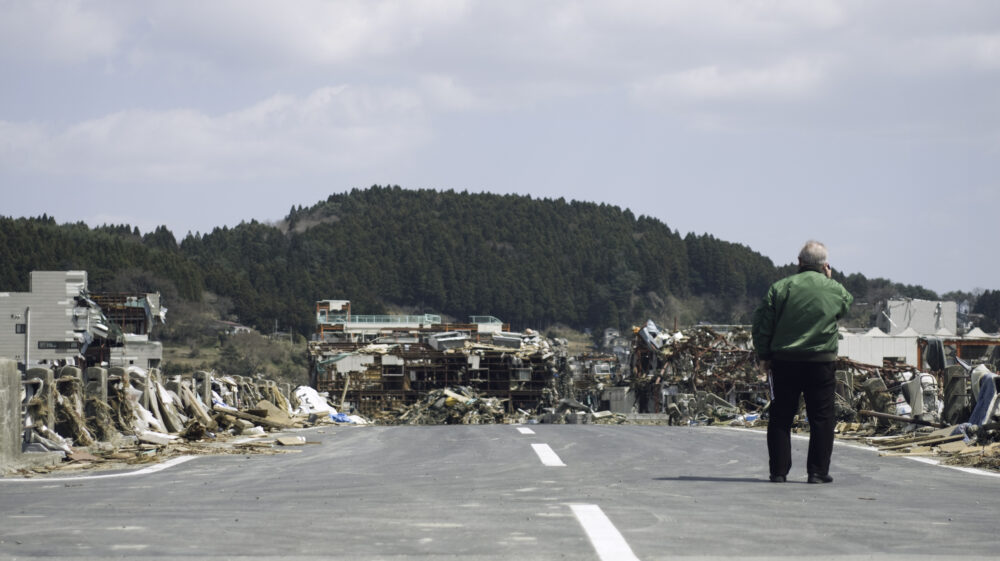 Older Japanese man in green jacket and black pants on phone in middle of road blocked by destroyed homes with tree-lined hill in background. Minami Sanrikucho devastated by earthquake and tsunami that struck Tohoku region of Japan on 11 March 2011, Miyagi-Ken
