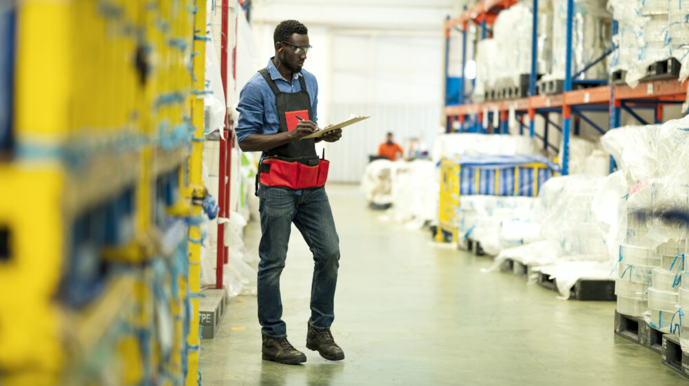 Black male worker wearing work apron and goggles holding clipboard and pen standing in large warehouse while counting inventory of material stock in factory