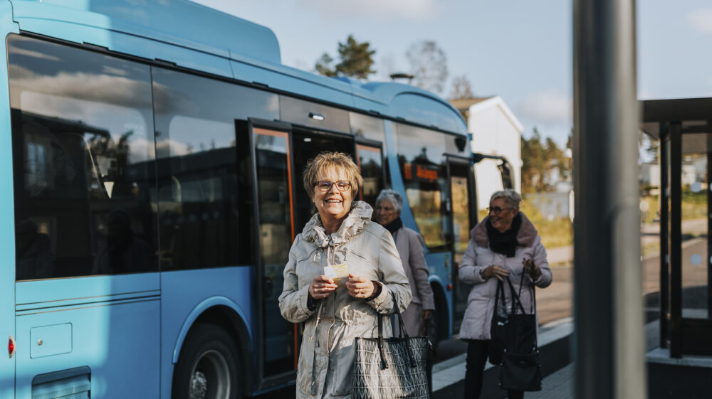 Smiling older woman wearing glasses at bus station standing in front of a blue bus holding a ticket in her hands, Sweden