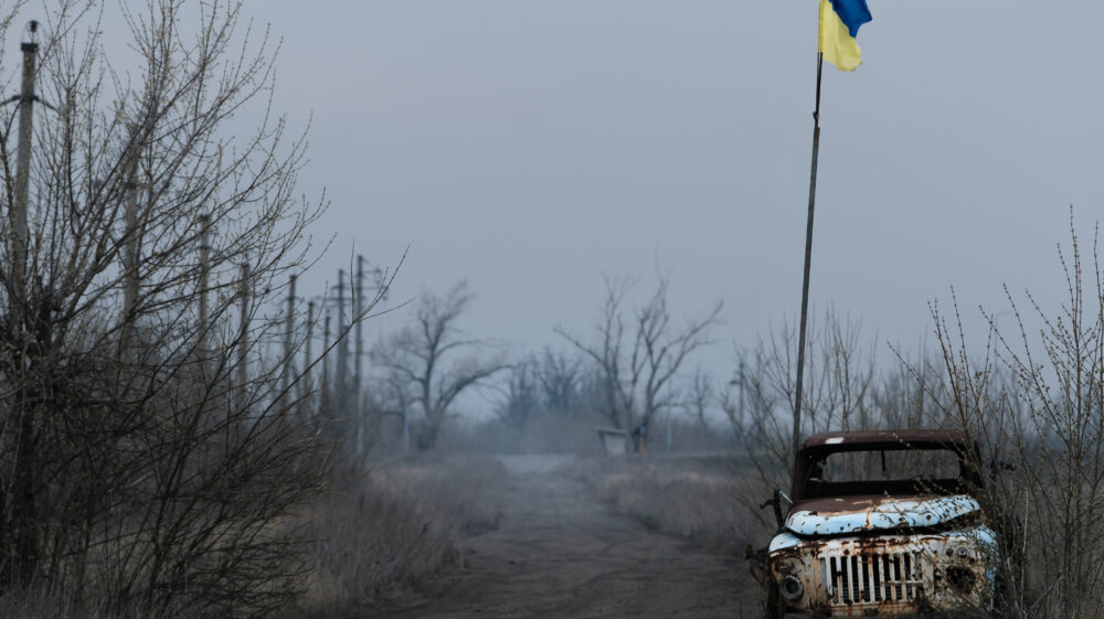 Grey empty landscape with leafless trees, rusty old white truck stands on side of muddy road next to Ukrainian flag on tall flagpole