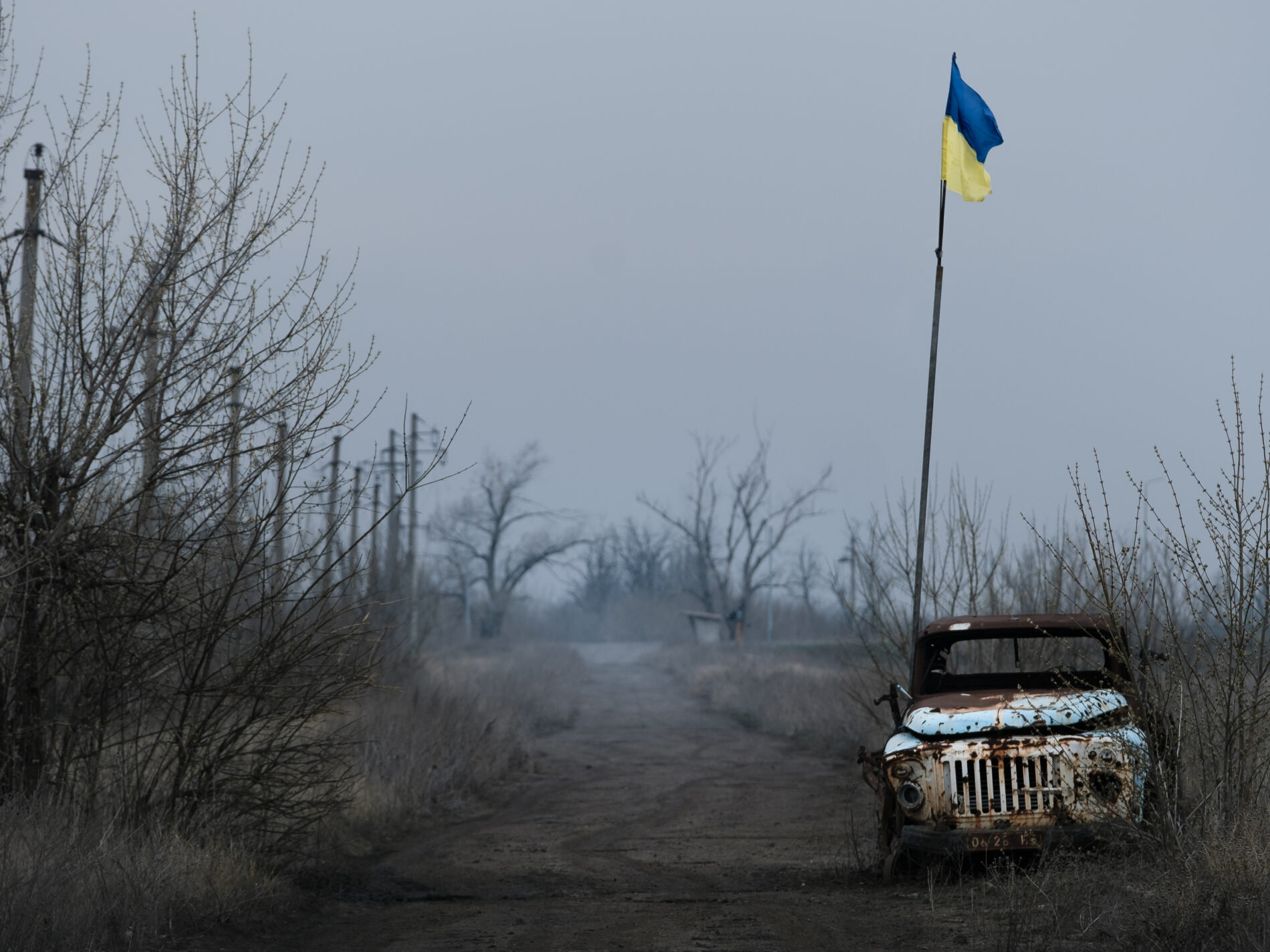 Grey empty landscape with leafless trees, rusty old white truck stands on side of muddy road next to Ukrainian flag on tall flagpole
