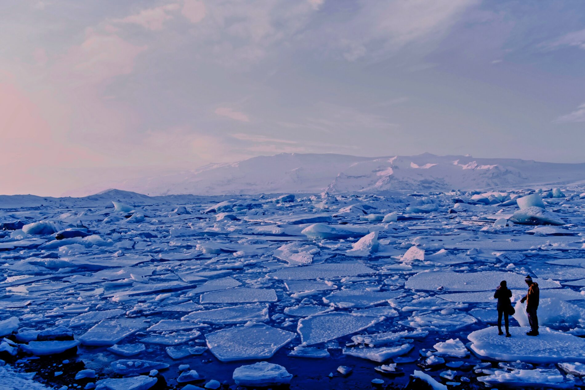 Man and woman stand on cracked sea ice beneath a gray sky, in sea of breaking ice sheets, Jökulsárlón, Iceland