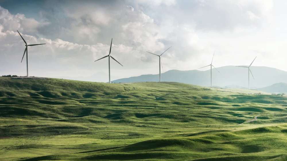 Green rolling hills and several wind turbines against a backdrop of mountains and clouds.