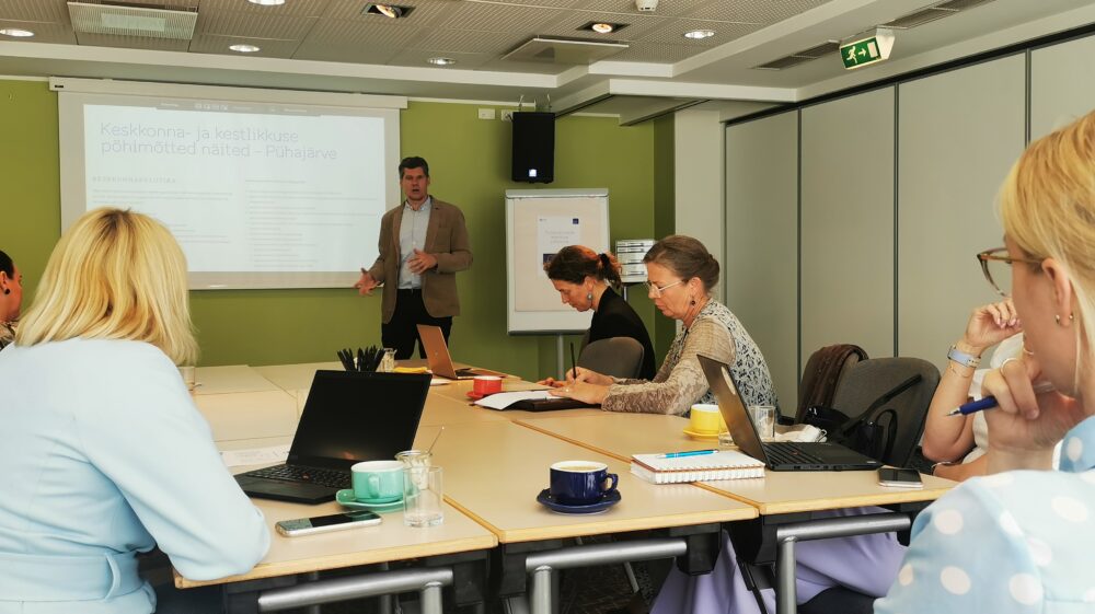 People seated around the table, looking at computers, a man givinb a presentation, standing in front of a screen.