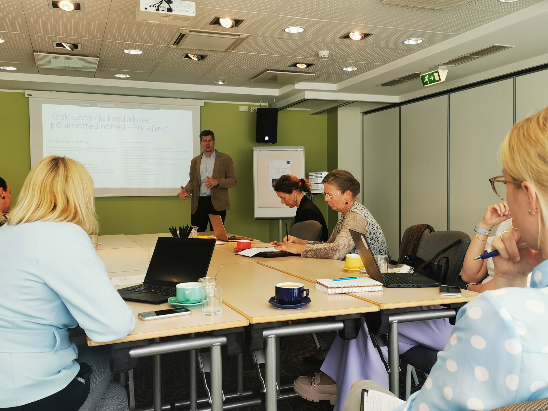 People seated around the table, looking at computers, a man givinb a presentation, standing in front of a screen.