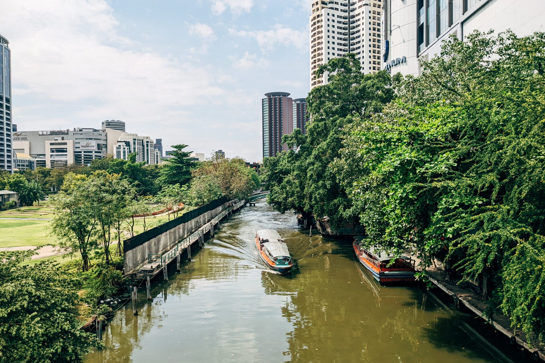 Waterway with boats between tree-lined high-rises and green grassy park with commercial buildings and high-rises in background, Bangkok, Thailand
