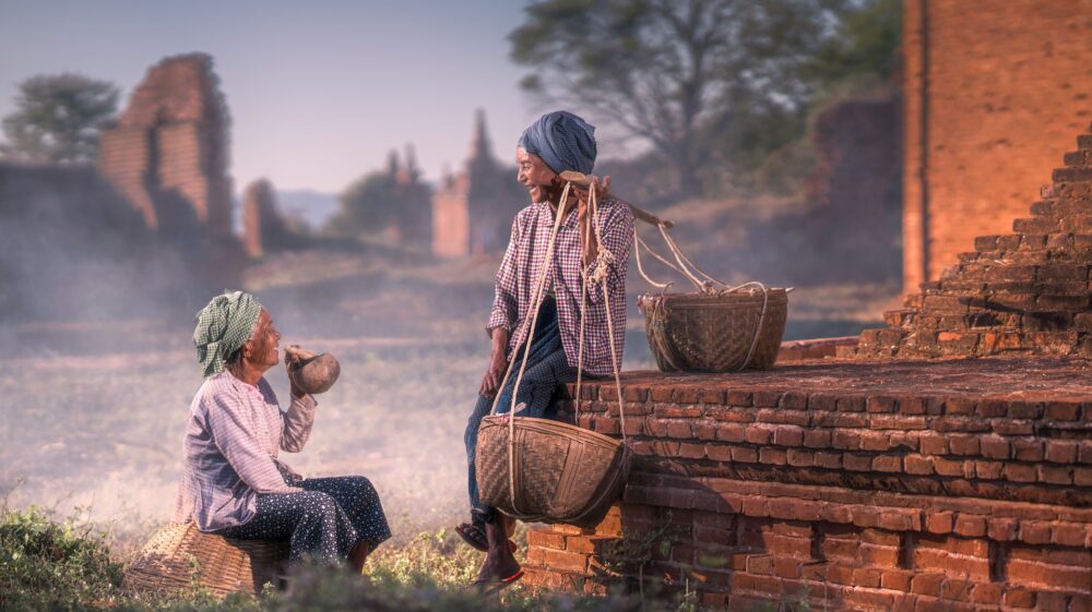 Two elderly ladies in traditional dress wearing headscarves chatting and having a laugh, one resting while carrying wicker baskets hanging off a pole, the other drinking a beverage with a wooden straw out of a gourd during early morning, Old Bagan, Myanmar