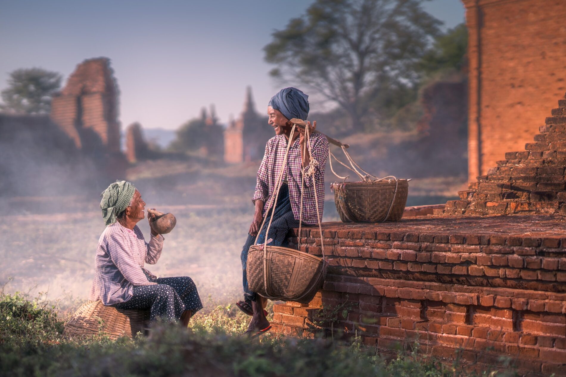 Two elderly ladies in traditional dress wearing headscarves chatting and having a laugh, one resting while carrying wicker baskets hanging off a pole, the other drinking a beverage with a wooden straw out of a gourd during early morning, Old Bagan, Myanmar