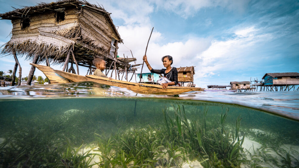 Bajau woman rowing wooden canoe in floating village carrying her child, showing the ocean floor and the wooden floating houses at the background, with blue sky and white clouds, Semporna, Sabah, Malaysia