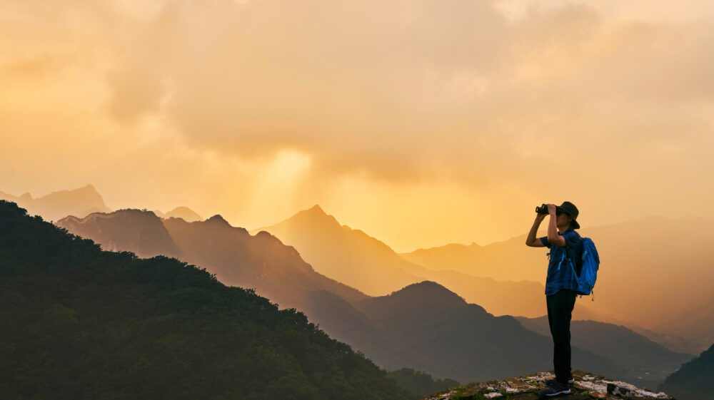 A man standing in the front of the picture, wearing a hat and a backpack, looking through binoculars. The background is colored orange, sun simmering through warm clouds. Under the orange sky stand several shilouettes of mountains, each one increasingly lighter and soaked in sun.