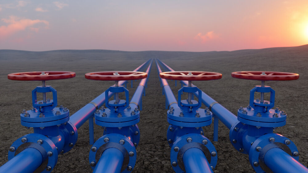 Red and blue gas or oil pipeline valves contrast with an expanse of brown ground against a pink sunset