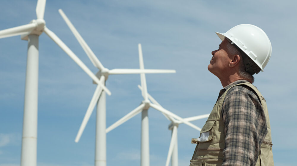 Man in white hard hat looks up at a row of wind turbines set against a blue sky filled with thin white clouds
