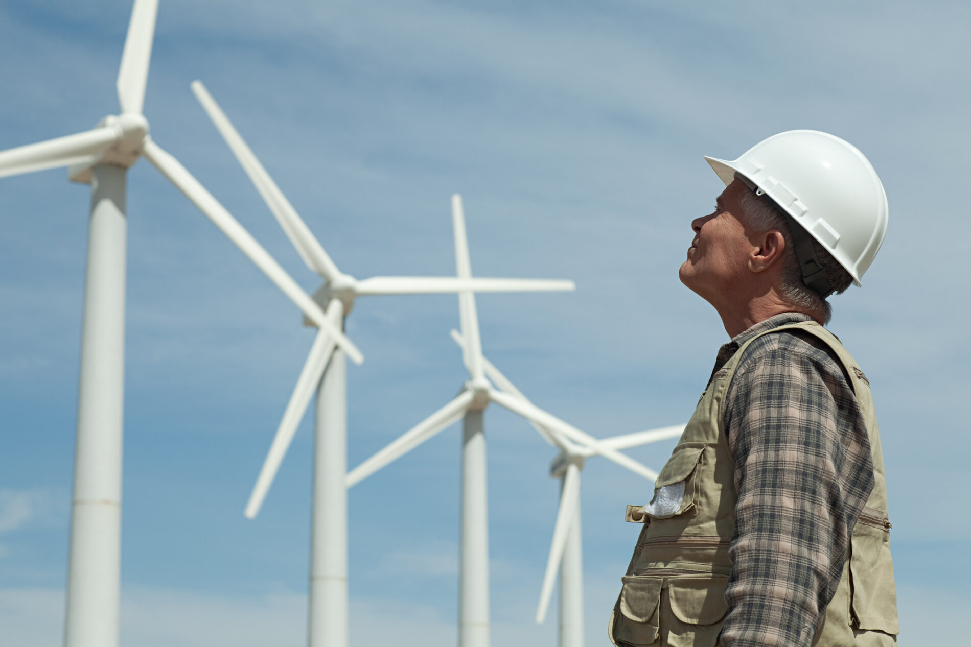 Man in white hard hat looks up at a row of wind turbines set against a blue sky filled with thin white clouds