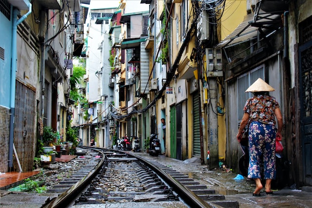 Older woman in straw hat and floral print shirt and pants and flip flops walking next to railway in crowded neighbourhood between high-rises on rainy day, Hanoi, Viet Nam