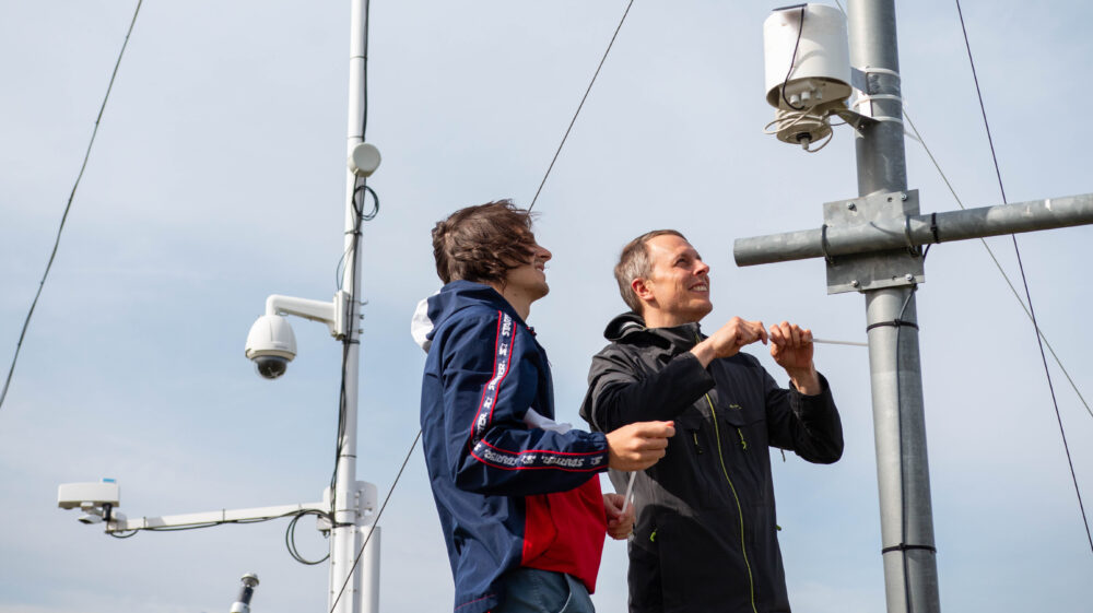 SEI Tallinn's intern Emile Neimry and senior expert Andreas Hoy installing a weather sensor. The sensor is a head-sized white gadget, which has been attached to a metal pole with several other poles and gadgets on the background.