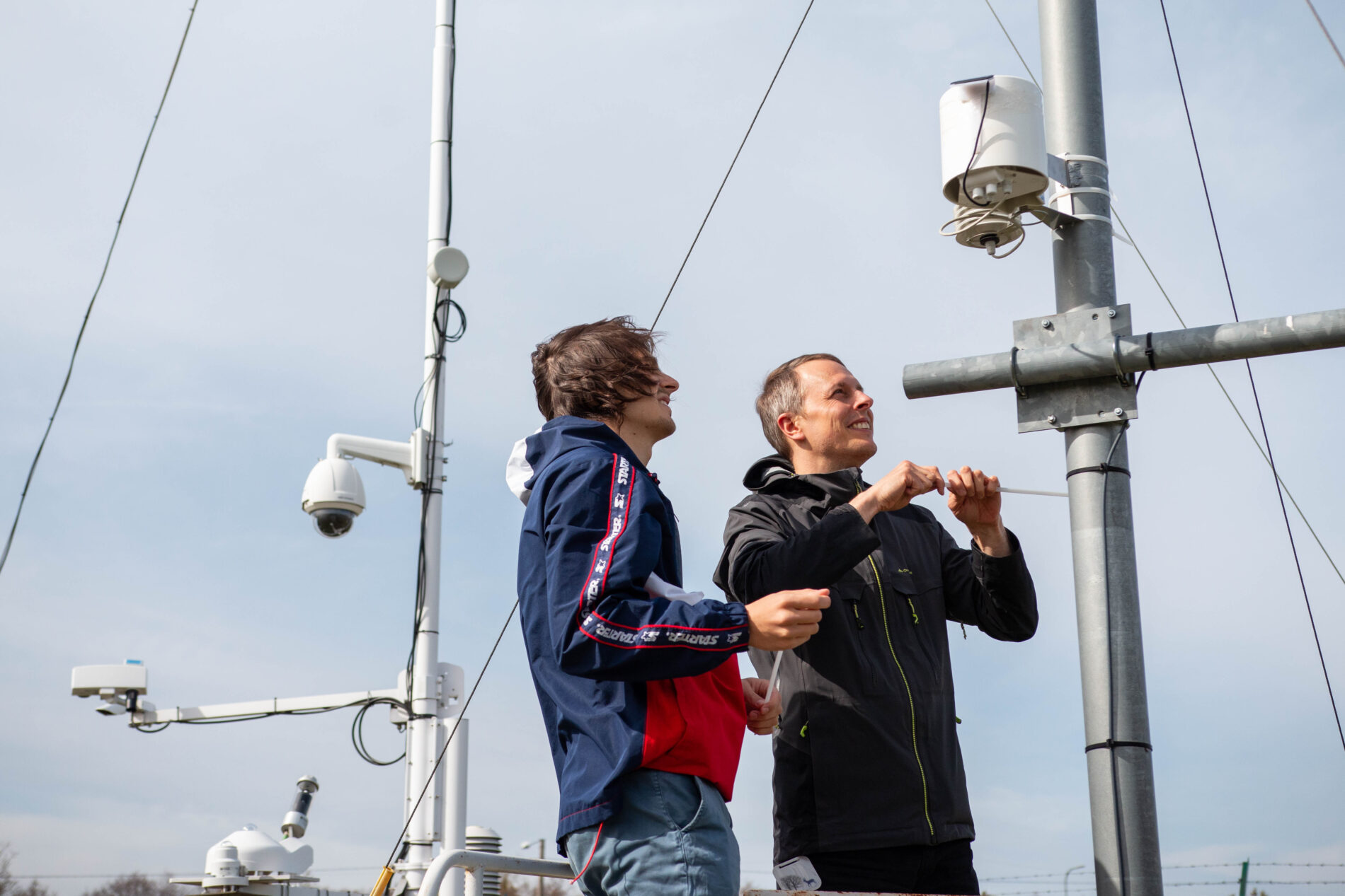 SEI Tallinn's intern Emile Neimry and senior expert Andreas Hoy installing a weather sensor. The sensor is a head-sized white gadget, which has been attached to a metal pole with several other poles and gadgets on the background.