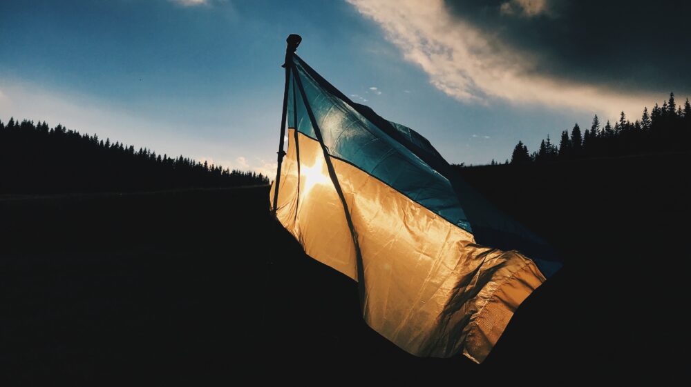 Ukrainian flag in foreground during daylight in forested area under blue sky with clouds, Ivano-Frankivs'ka oblast, Ukraine