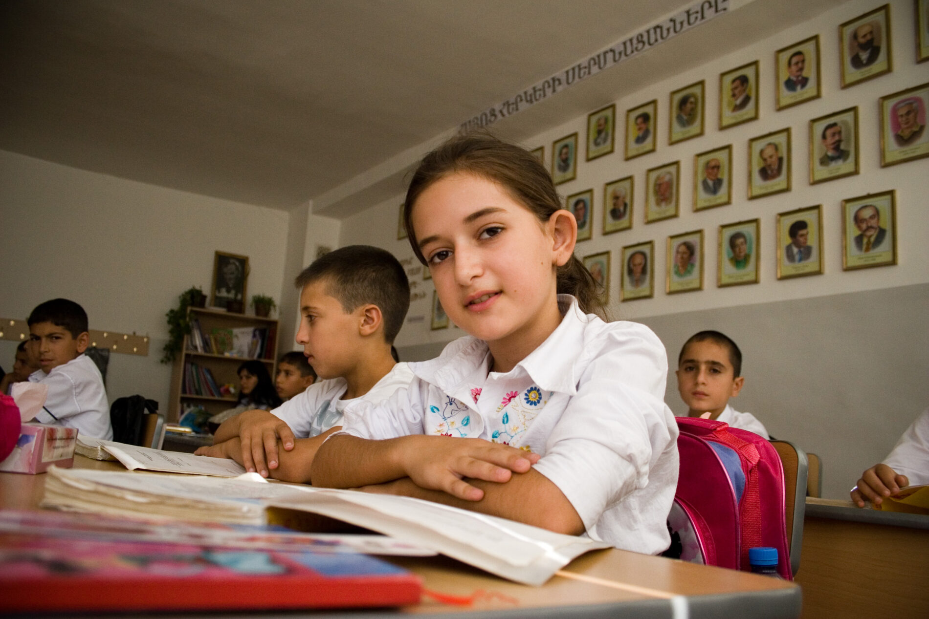 Children in a classroom, with a girl with folded arms looking at the camera and rows of portraits (men in profile) on the wall behind the children's desks. https://flickr.com/photos/asiandevelopmentbank/8429389750/