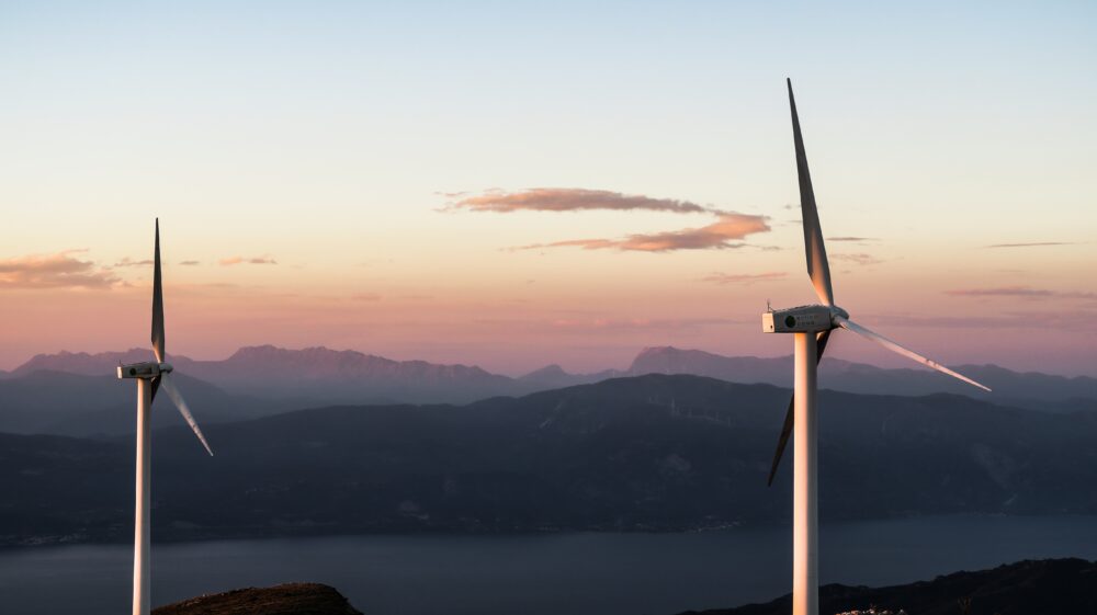 Two white wind turbines set in Greek mountains overlook a body of water.