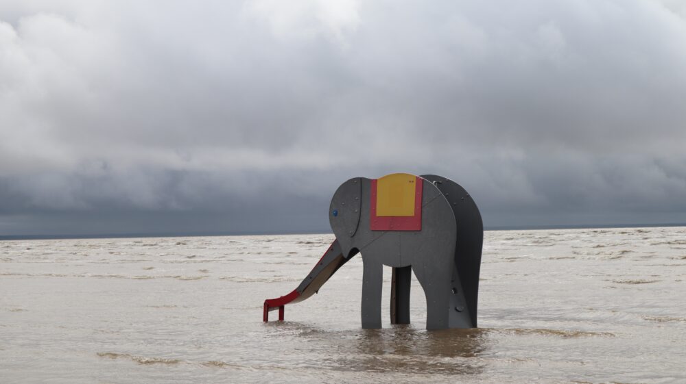 A construction of a grey wooden elephant slide standing in the Pärnu sea. The weather is grey and the sky is filled with clouds.