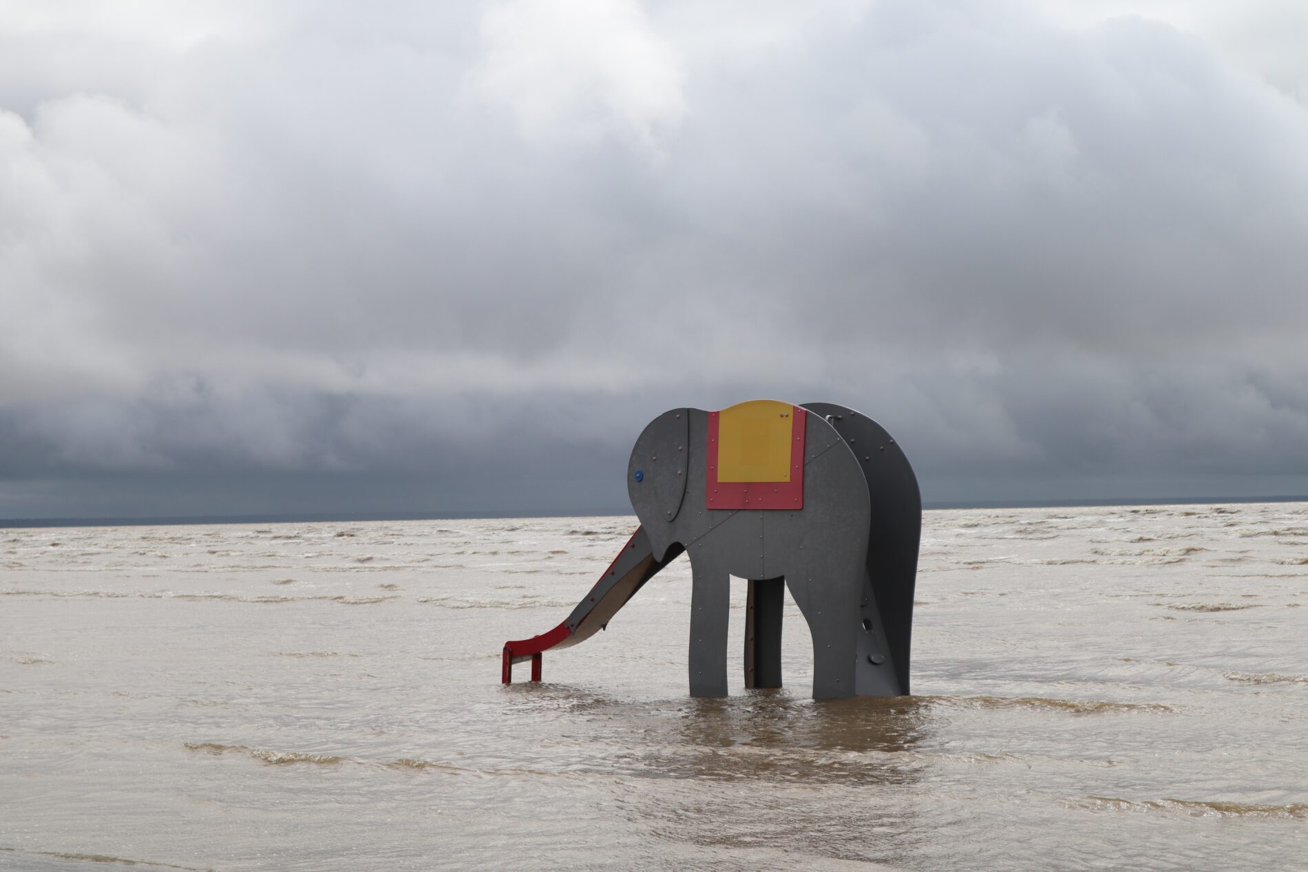 A construction of a grey wooden elephant slide standing in the Pärnu sea. The weather is grey and the sky is filled with clouds.
