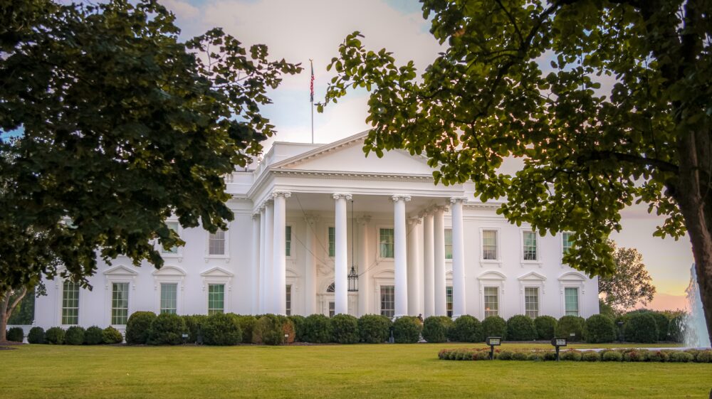 The White House in Washington D.C. is framed by leafy trees, foregrounded by a manicured green lawn