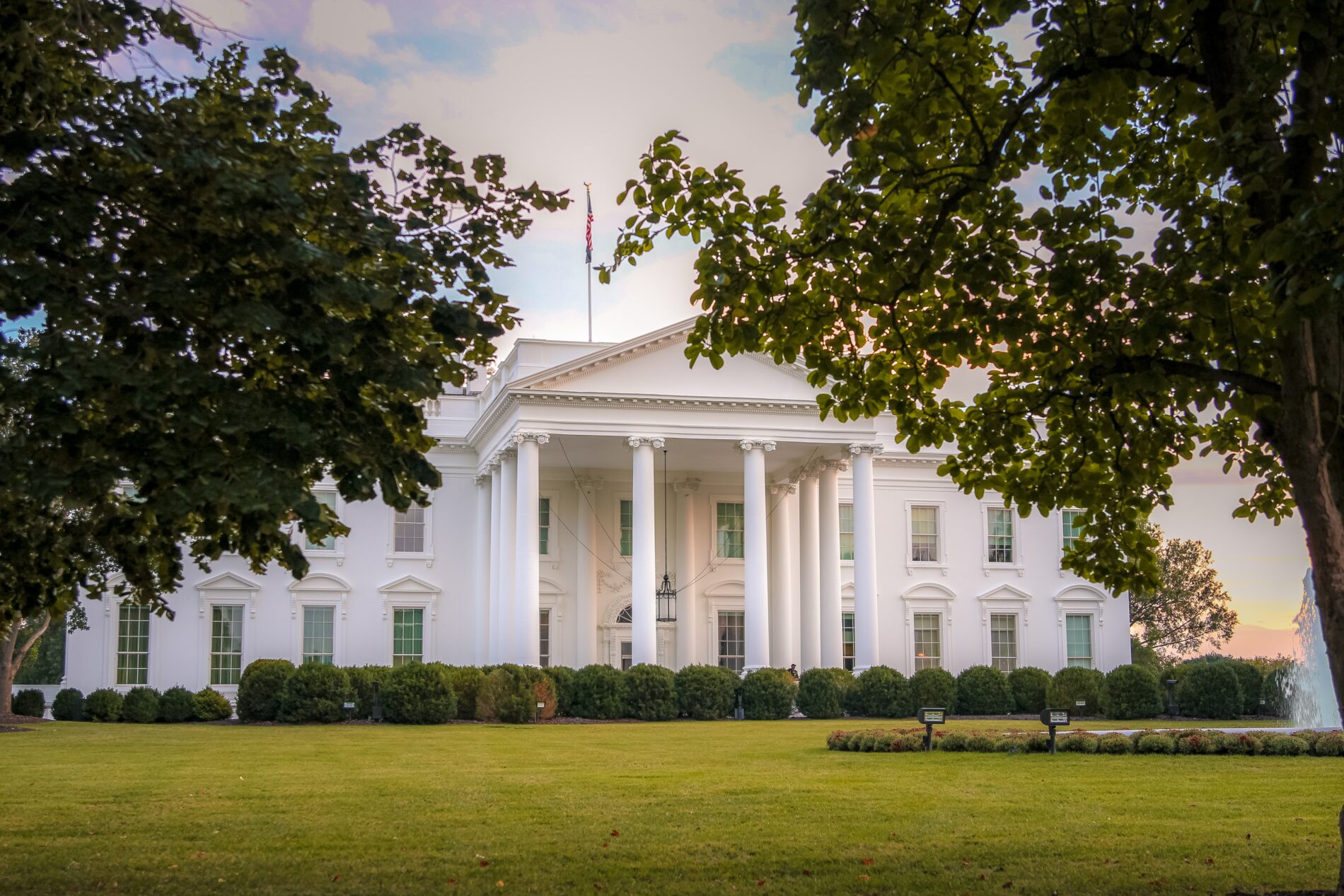 The White House in Washington D.C. is framed by leafy trees, foregrounded by a manicured green lawn