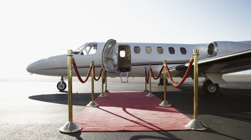 A red carpet flanked by velvet ropes leads to the entry of a private jet on a runway.