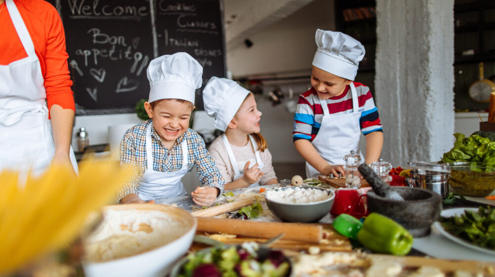 Three children with white chef hats cooking and laughing behind a table full of food on cooking supplies.