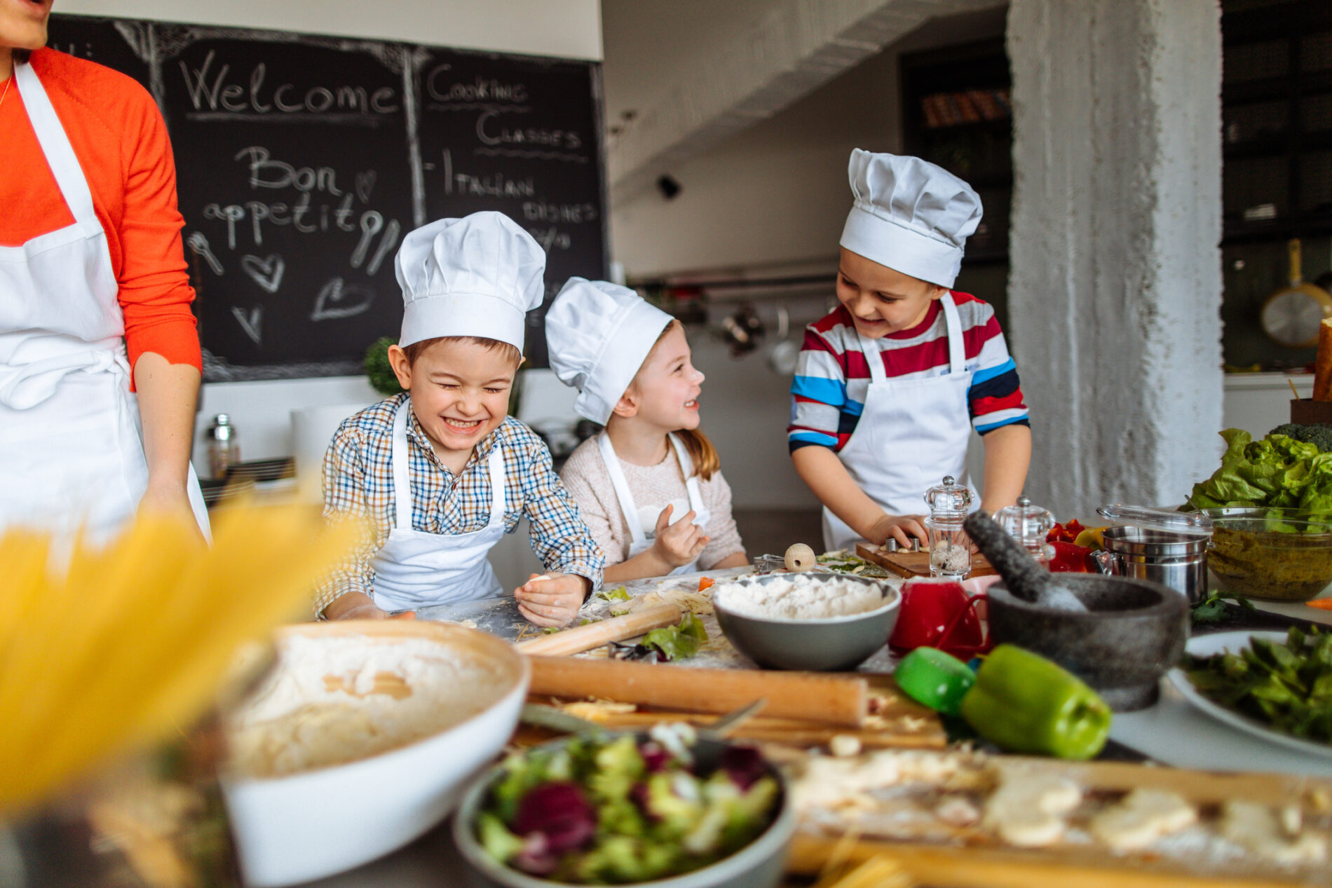 Three children with white chef hats cooking and laughing behind a table full of food on cooking supplies.