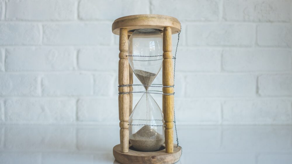 Brown sand pours through an hourglass with a light wooden outer frame in front of a white-painted brick wall