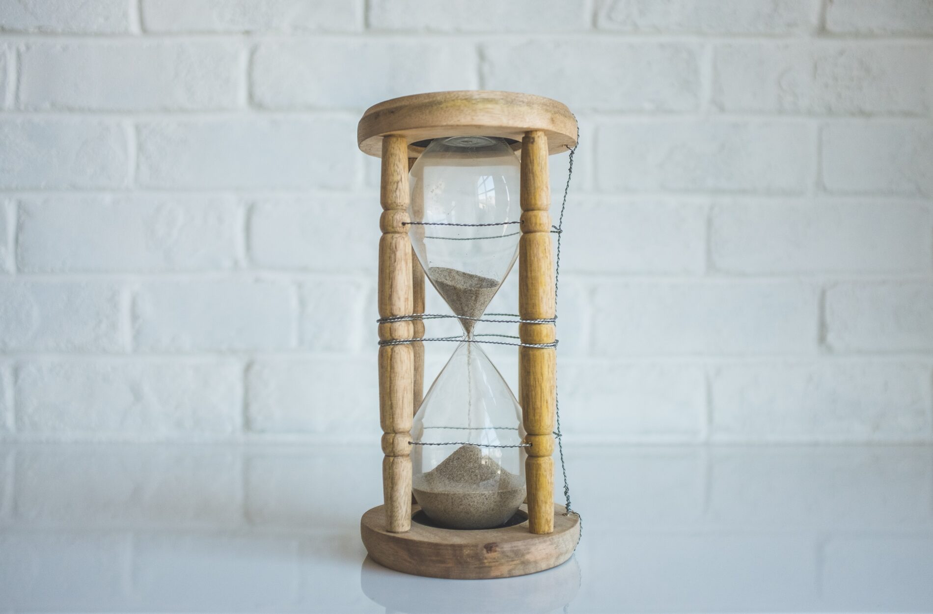 Brown sand pours through an hourglass with a light wooden outer frame in front of a white-painted brick wall