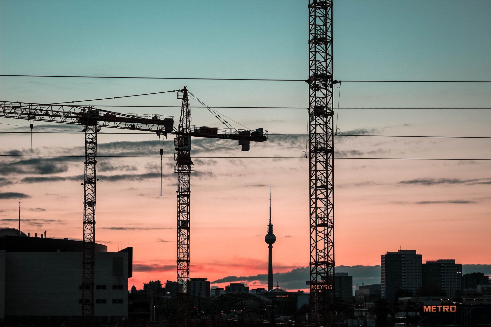 Construction cranes and a TV tower are silhouetted against an orange and blue sky in Berlin.