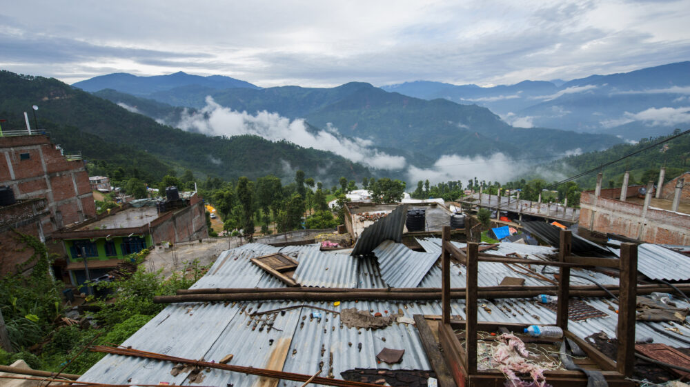 Buildings rattled by an earthquake in Nepal lie in ruins amid a summer monsoon, against a backdrop of lush forested mountains.