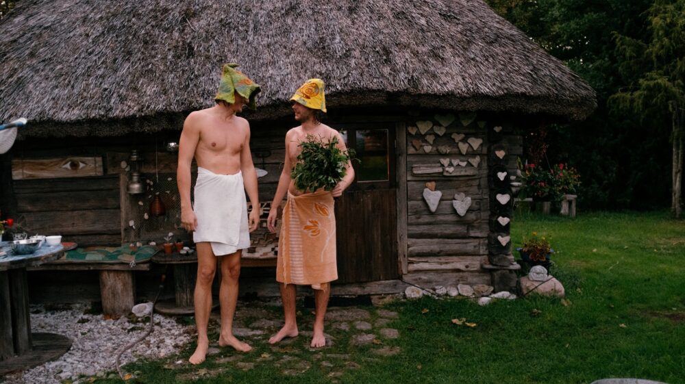 Two men standing in front of Estonian wooden sauna, wearing sauna hats, towels around their waist