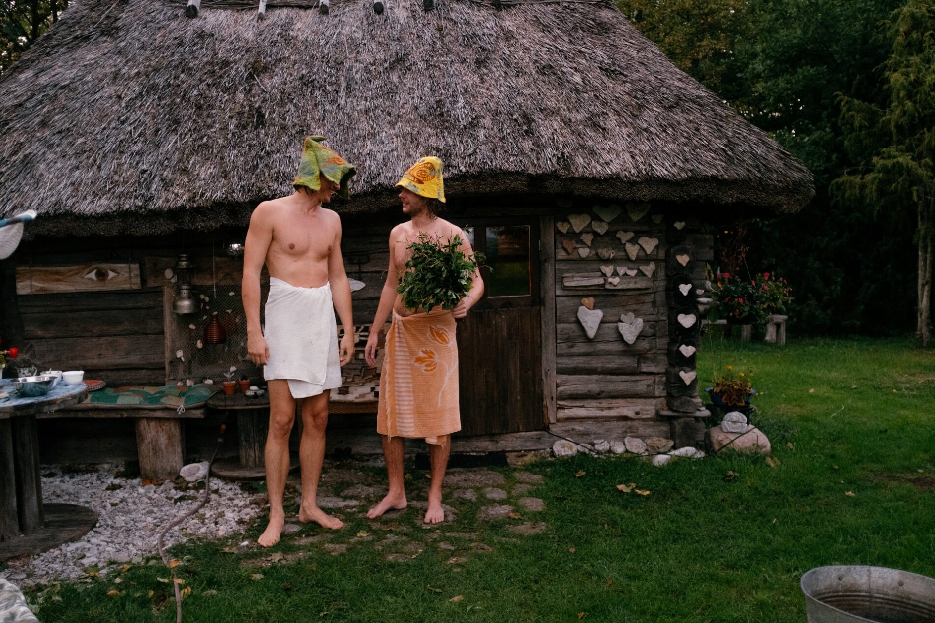 Two men standing in front of Estonian wooden sauna, wearing sauna hats, towels around their waist