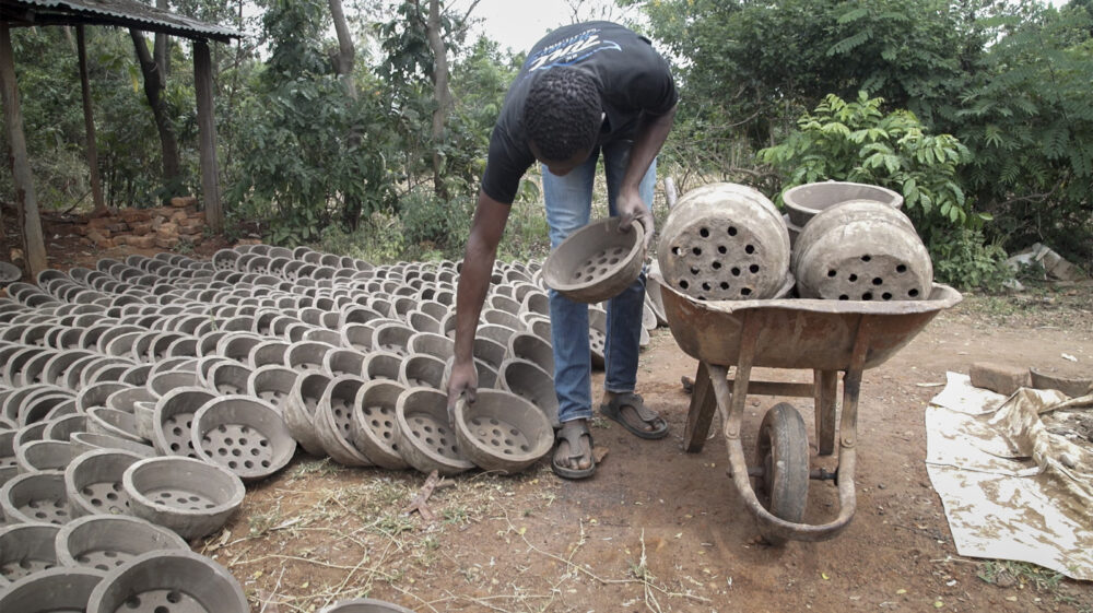 Person arranging rows of clay cooking stoves beside a wheelbarrow outdoors.