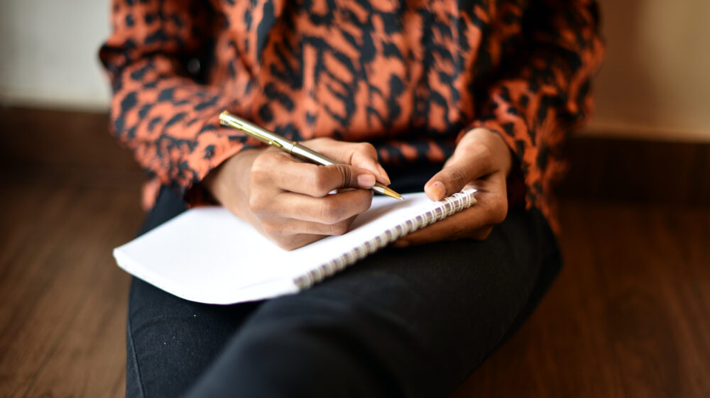 Close up of a woman's hands holding a pen writing on a notepad. She is sitting on the floor, leaning the notepad on her outstretched legs.