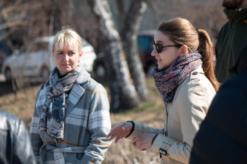 Two women: one with sunglasses is explaining and gesturing with hands, another is listening in the back.