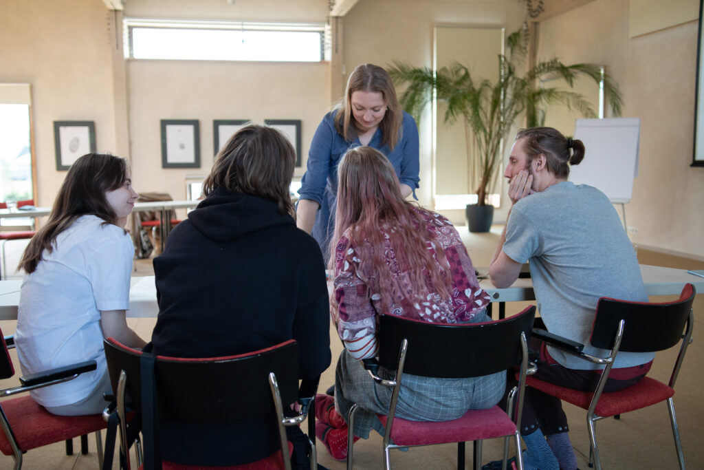 A woman placing something on a table before four young people who are photographed from the back. They are sitting on chairs in a big light room.