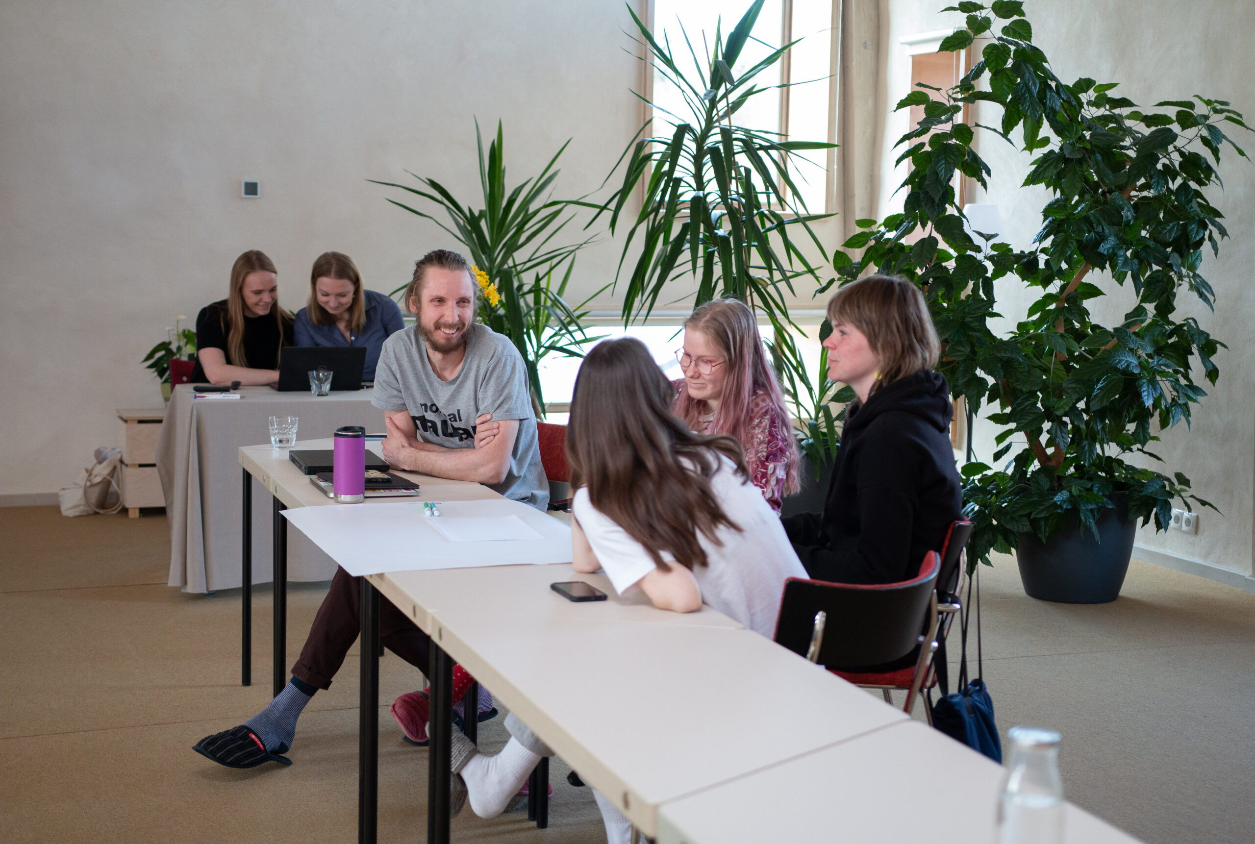 Four young people sitting behind a big table and discussing something, they are smiling. Behind is a smaller table with two women looking at something in the computer.