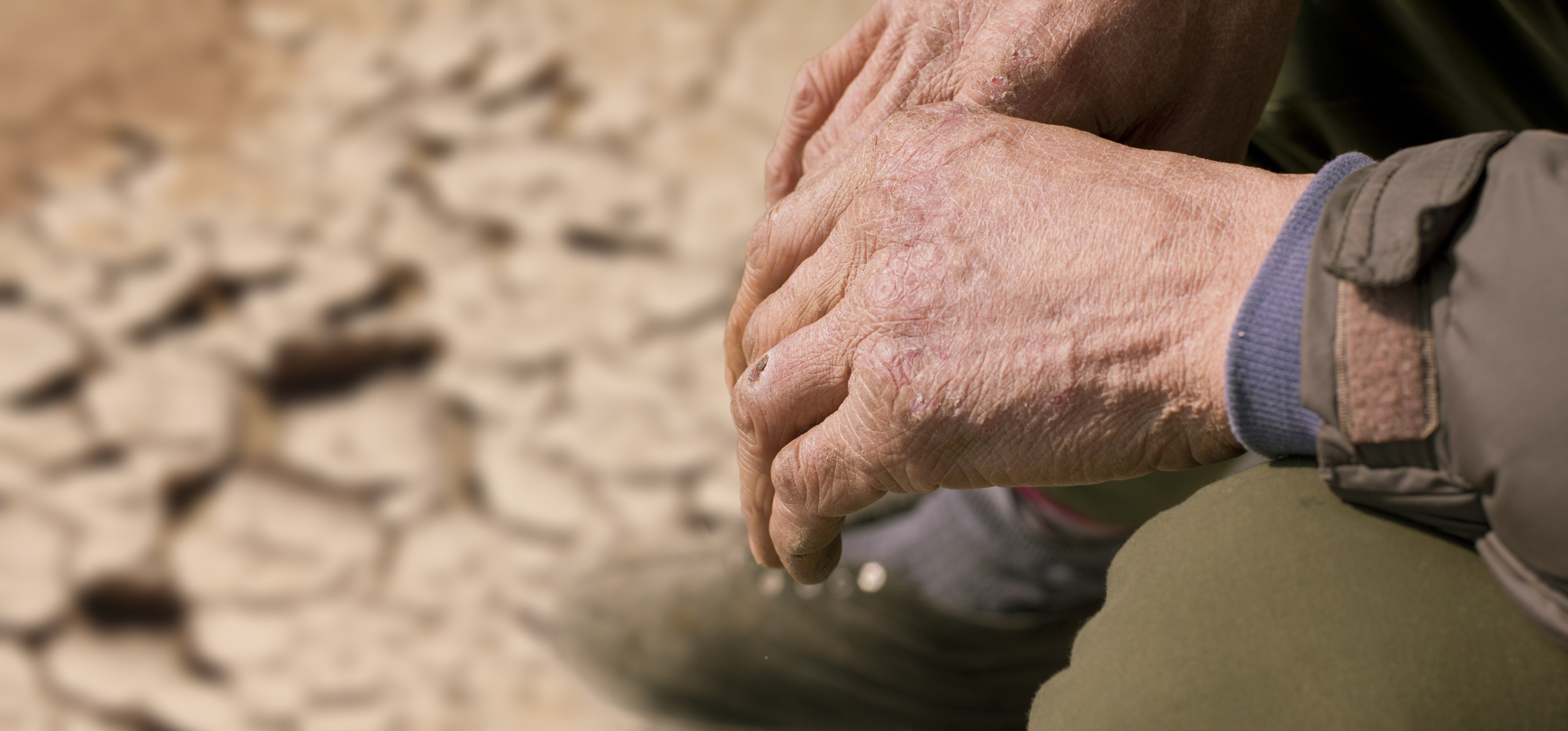 A close-up of hands of an older man sitting on a dried up field, cracks showing in the ground.
