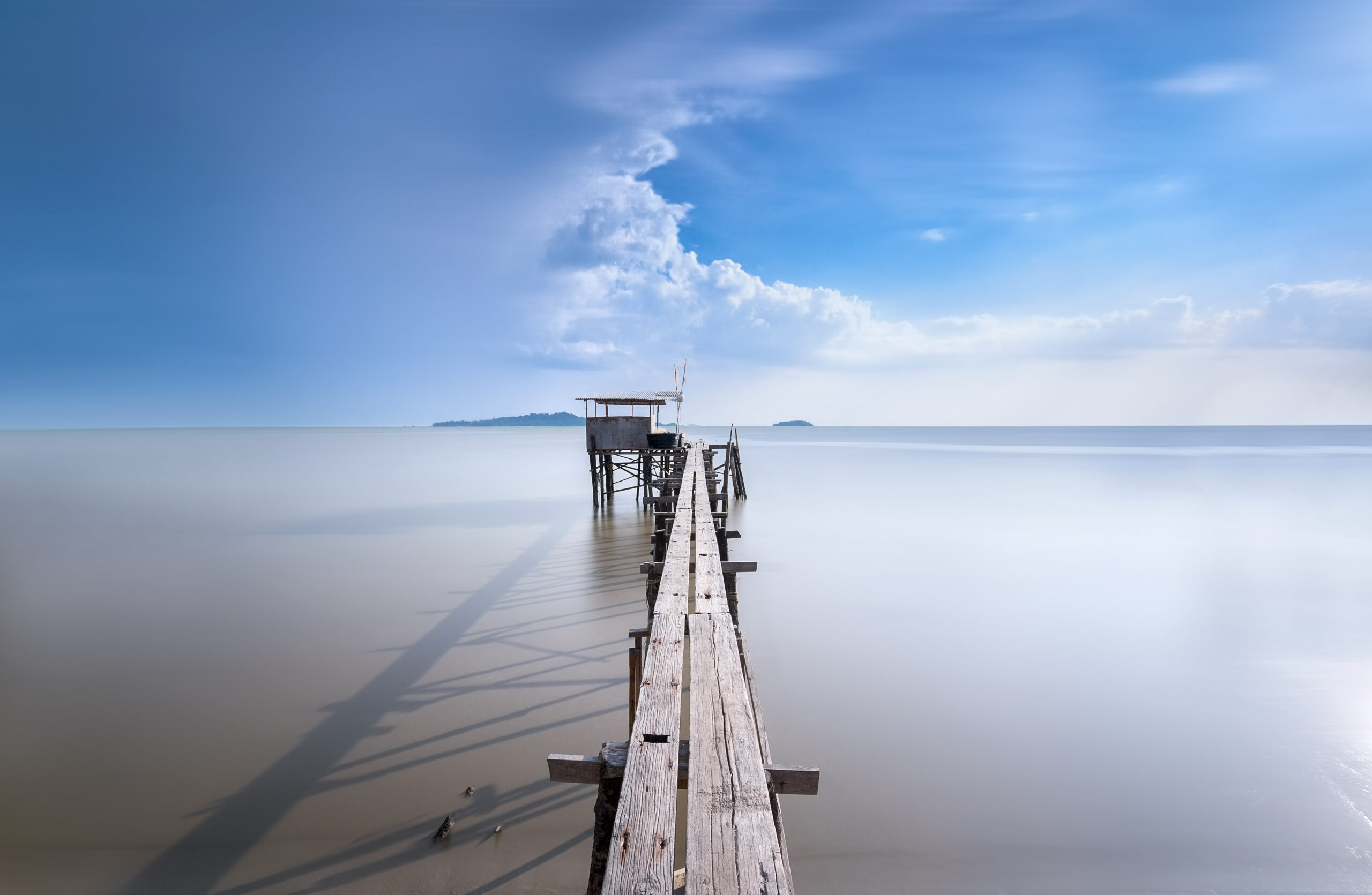 Wide landscape with a brown and blue colour palette, centred on an abadoned jetty which stretches out towards the horizon.