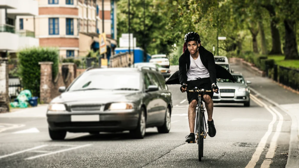A Black man cycles down a busy road as a car is moving to overtake him.