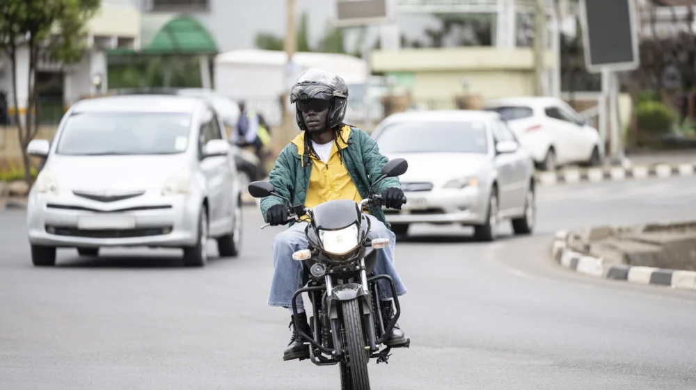 Wide shot of a man riding a motorbike on a road in Nairobi, with two cars in the background.