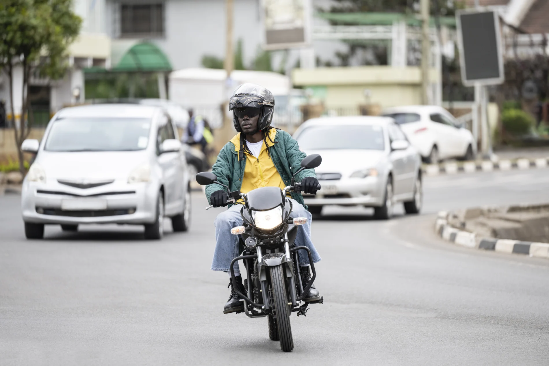Wide shot of a man riding a motorbike on a road in Nairobi, with two cars in the background.