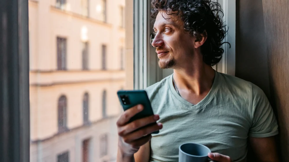 man drinking coffee and using smart phone while leaning on a window sill with Swedish building in the background.